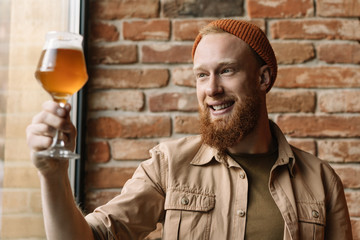 Portrait of young bearded hipster man with happy emotional face holding glass with craft beer, checking quality of alcohol, testing beer and smiling in loft brewery. Successful small business concept