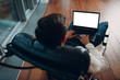 © primipil - A young man with laptop with blank white screen sits in a chair by the window in a skyscraper.