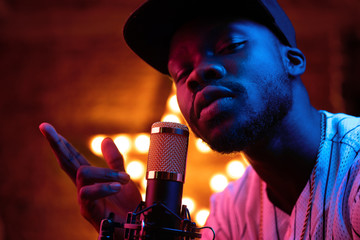 Young man with beard and mustaches in baseball cap and t-shirt sing in microphone and look in camera. Pink neon light