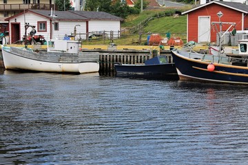 Fishing boats at the wharf, Petty Harbour, Newfoundland. Three boats tied up at the dock, two fishing sheds, crab traps on the pier, slight ripple on the water.