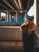 Girl With Long Hair In A Winter Jacket And Scarf Stands Under The Bridge At Sunset