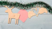 Tinsel With Christmas Ornaments And Blank Paper Note Wooden Background. Merry Christmas Concept. Christmas Ornaments And Wooden Deer And Heart Toy Shimmering Green Tinsel. Christmas Ornaments