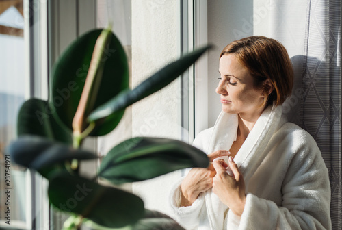 Cuadros en Lienzo A young woman with bath robe standing indoors by a window in the morning