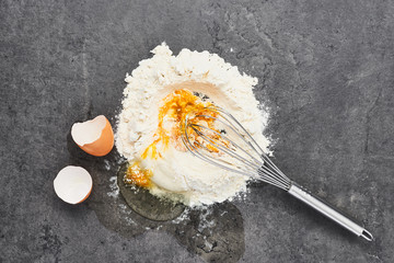 Mixing egg and flour. Egg, flour and whisk on dark grey concrete background. Ingredients for dough or noodles preparation. Top view. Copy space.