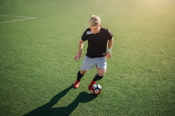 Young blonde football player playing on green lawn outside. He is going to kick ball.