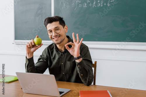 Fotografie, Obraz smiling male teacher in glasses holding apple and showing ok sign at computer de