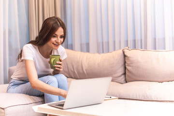 Happy woman is sitting on couch and typing on laptop. She holding green cup and looking at screen with smile. Copy space on right side