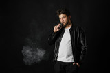 Fototapeta  - Man with vaping mod exhaling steam at black studio background. Bearded guy smoking e-cigarette to quit tobacco. Vapor and alternative nicotine free smoking concept