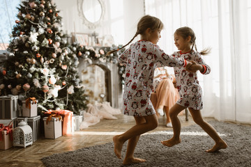 Two little sisters in pajamas having fun New Year's tree with gifts in the light cozy room