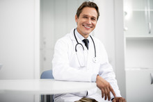 Emotional Confident Practitioner In White Coat Smiling And Putting One Hand On The Table While Sitting And Looking