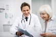© Yakobchuk Olena - Cheerful young doctor sitting close to his aged patient and smiling while holding clipboard and showing notes to the woman