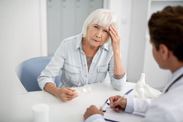 Wall Mural - Tired elderly woman visiting her practitioner and touching the forehead while having headache