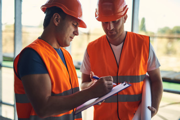 Calm concentrated foreman in the uniform holding clipboard and making notes on the paper while his colleague standing next to him