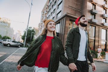 Waist up portrait of cheerful bearded man holding hand of his charming girlfriend. They looking away and smiling