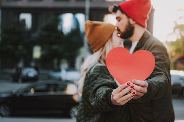 Close up of male and female hands holding sign of love. Bearded man in hat kissing forehead of his charming girlfriend