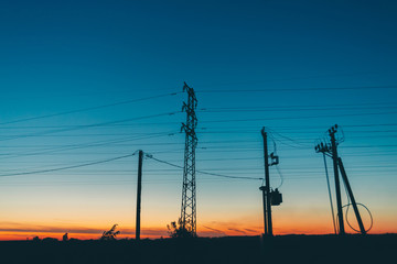 Power lines in field on sunrise background. Silhouettes of poles with wires at dawn. Cables of high voltage on warm orange blue sky. Power industry at sunset. Many cables in picturesque vivid sky.