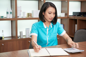 Pretty Asian woman working in spa salon and calculating while taking notes at working stand