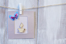 Card Of Gentle Lilac Color With Stylized Wooden Cup Of Coffee And Decorative Butterfly On Light Wooden Background.
