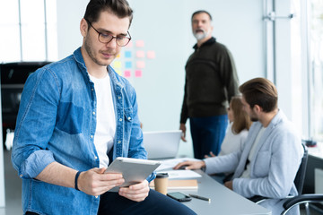 Canvas Print - Young and creative. Handsome young man holding digital tablet and smiling while his colleagues discussing something in the background.