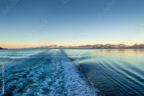 Fotografie, Obraz Ship wake at dawn, lighting rugged mountains across ocean channel