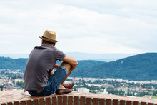 A Young Guy In Comfortable Moccasins Sits On The Edge Of A Tall Building And Looks At The Mountains And The City Of Graz