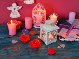 Valentine's Day decorative composition of burning candles in the form of roses, decorative lanterns with burning candles, red felt hearts, notebook and pencils, festive decor on a textural wooden tabl