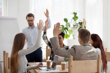 Diverse employees raise hands engaged in teambuilding activity during business mentor or coach flipchart presentation, multiethnic colleagues participate in education training during meeting