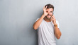 © Krakenimages.com - Handsome young man over grey grunge wall speaking on the phone with happy face smiling doing ok sign with hand on eye looking through fingers