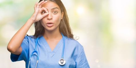 Wall Mural - Young arab doctor surgeon woman over isolated background doing ok gesture shocked with surprised face, eye looking through fingers. Unbelieving expression.