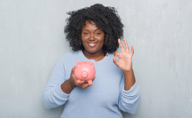 Sticker - Young african american woman over grey grunge wall holding piggy bank doing ok sign with fingers, excellent symbol