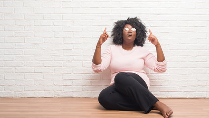 Poster - Young african american woman sitting on the floor at home amazed and surprised looking up and pointing with fingers and raised arms.