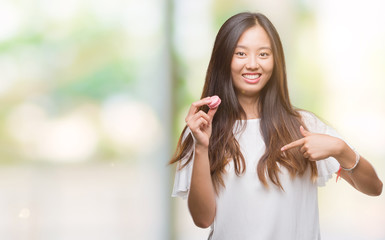 Canvas Print - Young asian woman eating pink macaron sweet over isolated background with surprise face pointing finger to himself