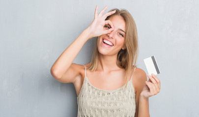 Wall Mural - Beautiful young woman over grunge grey wall holding credit card with happy face smiling doing ok sign with hand on eye looking through fingers