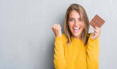 Wall Mural - Beautiful young woman over grunge grey wall eating chocolate bar screaming proud and celebrating victory and success very excited, cheering emotion