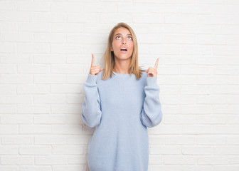 Wall Mural - Beautiful young woman over white brick wall amazed and surprised looking up and pointing with fingers and raised arms.