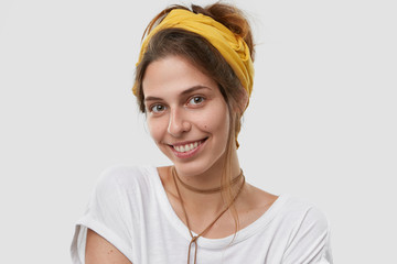 Wall Mural - Portrait of pleasant looking Caucasian woman with gentle smile on face, has dark combed hair, healthy skin, wears yellow headband, isolated over white background. Facial expressions concept.