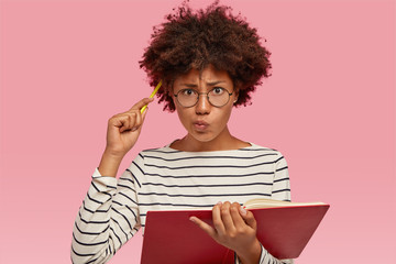 Indoor shot of confused indecisive woman with black skin, curly hair, scratches head with pencil, dressed in striped sweater, purses lips. holds diary, isolated over pink background. Hmm, let me think