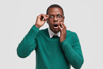 Wall Mural - Scared dark skinned guy keeps jaw dropped from fear, notices horrible scene in front, dressed in green jumper, wears eyewear, expresses surprisement and fear, models over white background. Emotions