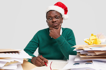 Photo of confused dark skinned thoughtful male, holds chin with hand, thinks about accounting report, writes down infromation in notepad, wears festive Santa Claus hat, busy with paper work at desktop