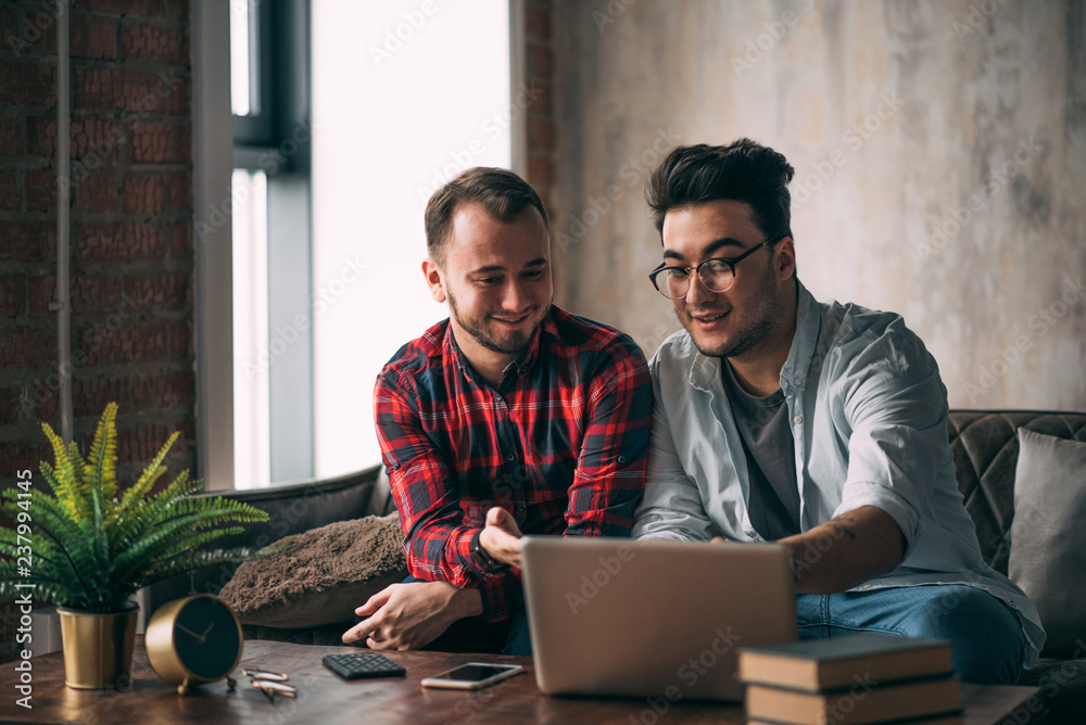 European same-sex male couple spend time together drinking coffee and watching laptop, sharing online interests , seeking for great ideas for their small business.