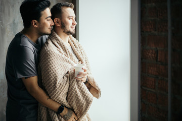 Wall Mural - Nice same-sex married couple is standing together at window. Careful and gentle male brunette embracing from the back his woman-like male friend wrapped in soft blanket. They are happy.