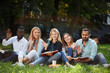 © alfa27 - Multiracial young friends during long break sitting in row at green grass lawn in the park, looking at camera, while welcoming with hand