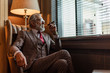 Serious old-aged businessman wearing classy custom tailored suit, red tie and expensive wristwatch sitting on old fashioned sofa in office, looking at camera and smoking cigar