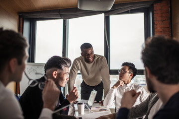 Canvas Print - deadline, technology and people concept - creative team of multiracial men, sitting round the desk in diverse casual and formal clothes talking over new project in office meeting room