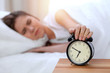 © rogerphoto - Alarm clock opposite of sleepy young woman stretching hand to ringing alarm willing turn it off. Early wake up, not getting enough sleep concept