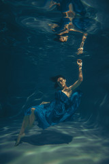 Wall Mural - elegant pretty woman swims underwater like a free diver in a blue evening dress alone