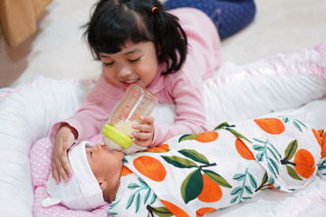 Wall Mural - Happy Asian toddler girl feeding her newborn baby sister a bottle of milk.