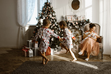 Wall Mural - Two little sisters in pajamas having fun New Year's tree with gifts in the light cozy room and their mother sits in the armchair with little baby next to the fireplace