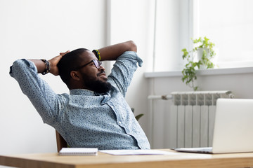 Black african millennial businessman take break during workday relaxing sitting on chair at office desk hold hands behind head looking at window thinking feels good. Stress relief daydreaming concept