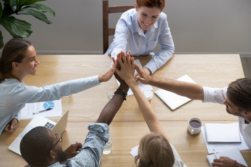 Diverse millennial partners company staff feels happy reached business goal giving high five stack palms together sitting at office desk in boardroom in conference, top above view. Team spirit concept